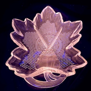 May include: A clear, leaf-shaped glass serving dish with a textured surface. The dish features a central circular design and a handle. The dish is likely used for serving snacks or small portions.