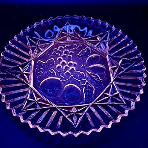May include: Clear glass serving dish with a starburst and geometric design. The center of the dish features an embossed design of grapes, leaves, and other fruit. The rim has a scalloped edge.