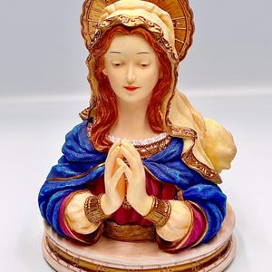 May include: A detailed bust of a religious figure with red hair, wearing a blue robe and a gold halo. The figure's hands are clasped in prayer. The base is a light beige color with gold accents.