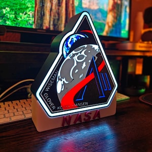May include: A triangular NASA-themed LED light with a black background and a white border. The design features a stylized Earth and moon, with the word "NASA" in red letters at the base. The names "Wiseman, Glover, Koch, Hansen" are also visible.