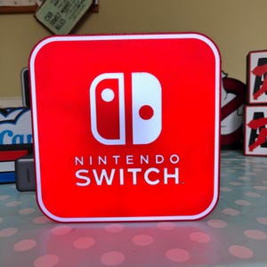 May include: A red square-shaped Nintendo Switch box with the Nintendo Switch logo in white. The logo features two vertical shapes, one with a white circle and the other with a white dot. The words "NINTENDO SWITCH" are printed below the logo in white.