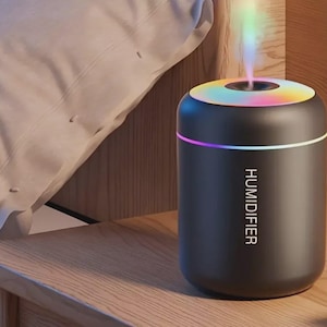 Op de afbeelding: Een zwarte luchtbevochtiger met een kleurrijke, verlichte bovenkant. Het woord "HUMIDIFIER" staat verticaal op de voorkant gedrukt. Het apparaat is cilindrisch en staat op een houten oppervlak, met een kussen op de achtergrond.