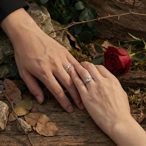 Puede incluir: Dos anillos de plata con diseños intrincados se muestran sobre una superficie de madera. Los anillos están en dos manos, con una rosa roja en el fondo. Los anillos parecen ser anillos de compromiso o de boda.