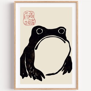 May include: A framed wall art print featuring a stylised black ink illustration of a frog against a light beige background. The frog has a large, rounded white belly and a red stamp-like seal with intricate characters in the upper left corner.