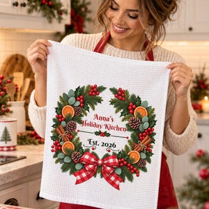 May include: A white waffle-weave kitchen towel featuring a printed holiday wreath made of pine branches, red berries, orange slices, pinecones, and cinnamon sticks. The text reads Anna's Holiday Kitchen and Est. 2026 above a red and white plaid bow.