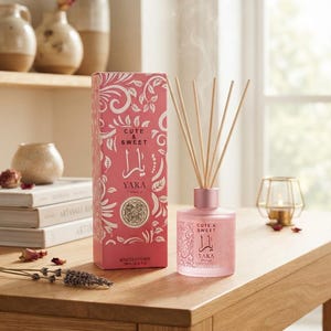 Könnte beinhalten: Ein rosafarbener Glas-Raumduft-Diffusor mit passender rechteckiger Schachtel, beide mit weißen Blumenmustern und dem Text Cute & Sweet Yara versehen. Der Diffusor enthält mehrere Holzstäbchen und ist mit 100ml / 3.4 fl. oz. beschriftet.