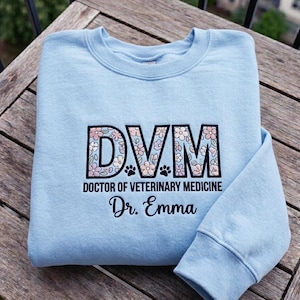 May include: Light blue sweatshirt with the letters "DVM" in a floral design, with the words "DOCTOR OF VETERINARY MEDICINE" and "Dr. Emma" printed below. The sweatshirt has paw prints under the letters.