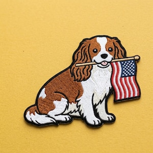 May include: Embroidered patch featuring a Cavalier King Charles Spaniel dog holding a small American flag in its mouth. The dog has brown and white fur with black outlines, set against a yellow background.
