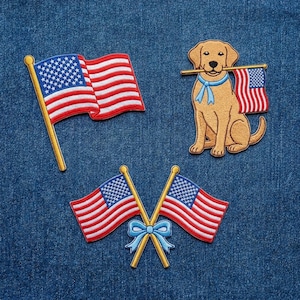 Może przedstawiać: Haftowane naszywki przedstawiające amerykańską flagę i psa golden retriever trzymającego flagę. Naszywki są czerwone, białe i niebieskie, ze złotymi akcentami. Naszywki znajdują się na niebieskim tle z dżinsu.