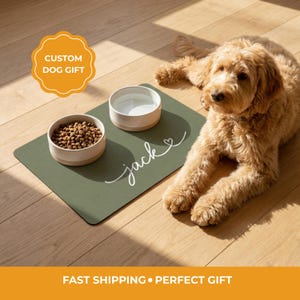 Peut inclure: Un tapis pour animal de compagnie vert olive rectangulaire avec le prénom Jack écrit en blanc en cursive et une petite icône de cœur. Deux bols ronds en céramique à intérieur blanc et base beige sont posés dessus, l'un rempli de croquettes.