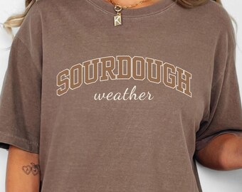 May include: Brown t-shirt with the text "SOURDOUGH weather" in a collegiate style font. The shirt is paired with denim shorts. A clear glass with a straw and a light brown beverage is held in the hand.