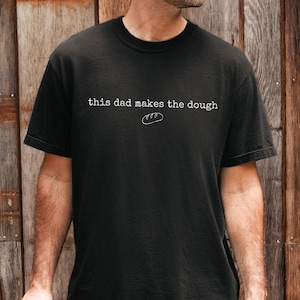 May include: Black t-shirt with the text "this dad makes the dough" and a bread graphic. The shirt is made of a soft material and is a casual wear item.
