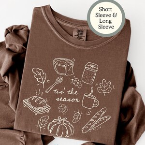 May include: An espresso-colored shirt featuring a white line-art graphic of a bowl, jar, spoon, leaves, bread, pumpkin, and mug with the text 'tis' the season'. A circular label indicates 'Short Sleeve & Long Sleeve' and the text 'Macy Dixie Designs' is visible.