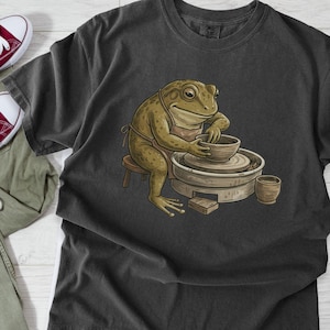 May include: Dark gray t-shirt featuring a cartoon frog crafting pottery. The frog is wearing an apron and sitting on a small stool. The shirt is paired with red sneakers and olive green pants.