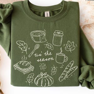 May include: A military green crewneck sweatshirt featuring a white line art graphic of a bowl, a jar, a spoon, a mug, a loaf of bread, a pumpkin, and leaves, with the text 'tis' the season' printed in the center of the design.