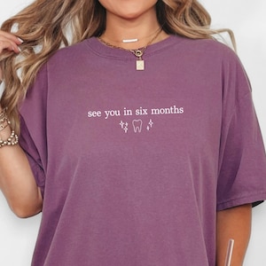 K&ouml;nnte beinhalten: Beerenfarbenes T-Shirt mit dem Text "see you in six months" und einer Zahn-Grafik. Das Shirt hat kurze &Auml;rmel und einen Rundhalsausschnitt. Das Shirt ist einfarbig und hat eine lockere Passform.