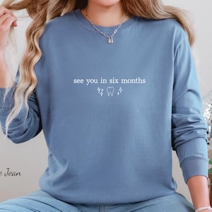 K&ouml;nnte beinhalten: Blaues Langarm-Sweatshirt mit dem Text "see you in six months" und einer Zahn-Grafik in Wei&szlig;. Das Sweatshirt hat eine einfarbige blaue Farbe. Der Text und die Grafik sind mittig auf der Brust platziert.