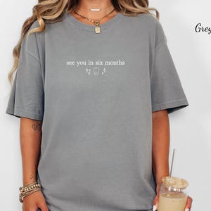 K&ouml;nnte beinhalten: Ein graues T-Shirt mit dem Text "see you in six months" und zahnmedizinischen Grafiken. Das Shirt hat eine lockere Passform und scheint aus weichem Material zu sein. Das Bild zeigt auch eine Person, die Schmuck tr&auml;gt und ein Getr&auml;nk h&auml;lt.