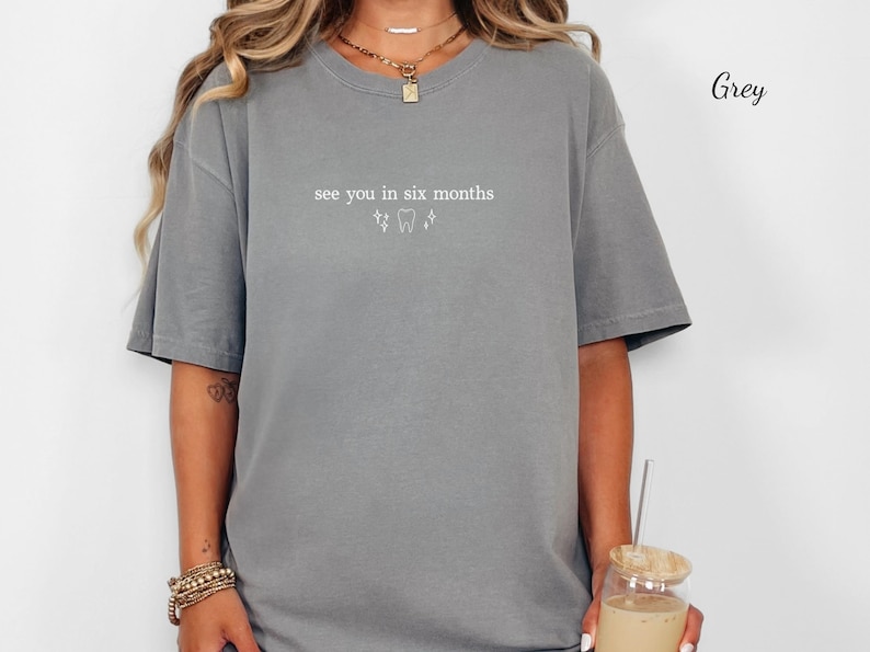 K&ouml;nnte beinhalten: Ein graues T-Shirt mit dem Text "see you in six months" und zahnmedizinischen Grafiken. Das Shirt hat eine lockere Passform und scheint aus weichem Material zu sein. Das Bild zeigt auch eine Person, die Schmuck tr&auml;gt und ein Getr&auml;nk h&auml;lt.
