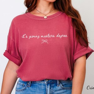 Puede incluir: Camiseta de color carmes&iacute; con mangas enrolladas. La camiseta presenta la frase "it's giving masters degree" en escritura blanca, con un peque&ntilde;o lazo gr&aacute;fico debajo. La camiseta se combina con unos vaqueros azules.