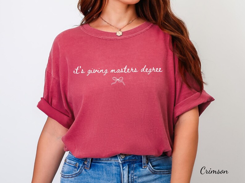 Puede incluir: Camiseta de color carmes&iacute; con mangas enrolladas. La camiseta presenta la frase "it's giving masters degree" en escritura blanca, con un peque&ntilde;o lazo gr&aacute;fico debajo. La camiseta se combina con unos vaqueros azules.