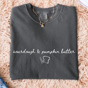 May include: A charcoal gray crewneck t-shirt featuring the white cursive text sourdough & pumpkin butter above a simple white line drawing of a butter knife and a slice of bread. A gold-toned chain necklace rests on the collar of the shirt.