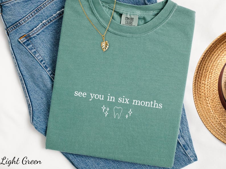 K&ouml;nnte beinhalten: Hellgr&uuml;nes T-Shirt mit dem Text "see you in six months" und einer Zahngrafik. Das Shirt wird auf einer blauen Jeans mit einer goldenen Halskette und einem Strohhut pr&auml;sentiert.