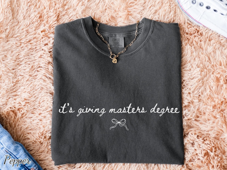 Puede incluir: Camiseta gris oscuro con la frase "it's giving masters degree" en escritura blanca y un lazo. Un collar de cadena dorada con un colgante descansa sobre la camiseta. La camiseta est&aacute; sobre una superficie suave color melocot&oacute;n.