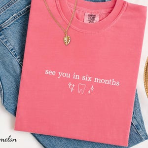 K&ouml;nnte beinhalten: Ein pinkfarbenes T-Shirt mit dem Text "see you in six months" und einer Zahngrafik. Eine goldene Halskette mit einem Blattanh&auml;nger und ein Strohhut sind ebenfalls zu sehen.