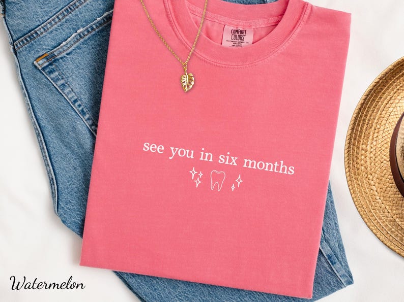 K&ouml;nnte beinhalten: Ein pinkfarbenes T-Shirt mit dem Text "see you in six months" und einer Zahngrafik. Eine goldene Halskette mit einem Blattanh&auml;nger und ein Strohhut sind ebenfalls zu sehen.