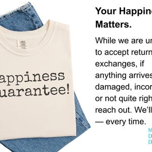 K&ouml;nnte beinhalten: Cremefarbenes T-Shirt mit dem Text "Happiness Guarantee!" in dunkelgrauen Buchstaben. Das Shirt ist auf einer blauen Jeans gefaltet. Der Text "Your Happiness Matters" ist ebenfalls sichtbar.