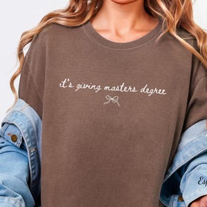 Puede incluir: Camiseta marr&oacute;n con la frase "it's giving masters degree" en escritura blanca y un lazo. La palabra "Espresso" est&aacute; escrita en la manga de una chaqueta vaquera.