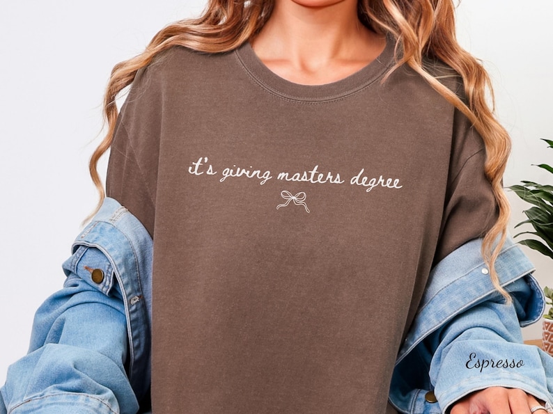 Puede incluir: Camiseta marr&oacute;n con la frase "it's giving masters degree" en escritura blanca y un lazo. La palabra "Espresso" est&aacute; escrita en la manga de una chaqueta vaquera.