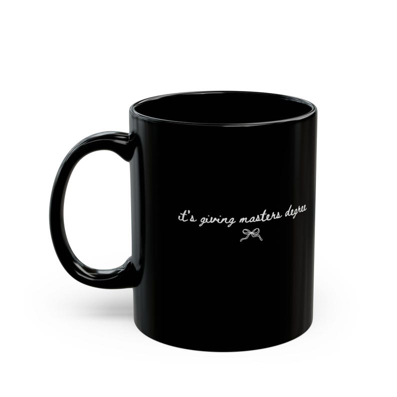 Puede incluir: Taza de caf&eacute; de cer&aacute;mica negra con asa curva. La taza presenta la frase "it's giving masters degree" en escritura blanca, acompa&ntilde;ada de un peque&ntilde;o lazo. Ideal para bebidas calientes.