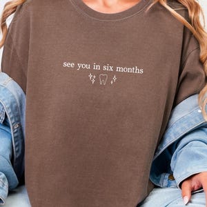 K&ouml;nnte beinhalten: Braunes T-Shirt mit dem wei&szlig;en Text "see you in six months", zusammen mit Zahn- und Sternsymbolen. Das l&auml;ssige Shirt ist einfarbig. Eine Jeansjacke wird &uuml;ber den Schultern getragen.