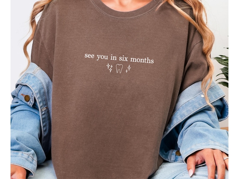 K&ouml;nnte beinhalten: Braunes T-Shirt mit dem wei&szlig;en Text "see you in six months", zusammen mit Zahn- und Sternsymbolen. Das l&auml;ssige Shirt ist einfarbig. Eine Jeansjacke wird &uuml;ber den Schultern getragen.