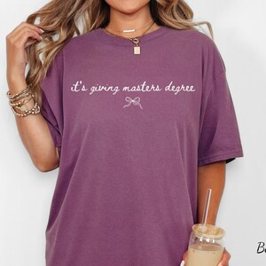 Puede incluir: Camiseta color bayas con la frase "it's giving masters degree" en escritura blanca. Un peque&ntilde;o lazo gr&aacute;fico est&aacute; debajo del texto. La camiseta es de corte holgado y tiene mangas cortas.