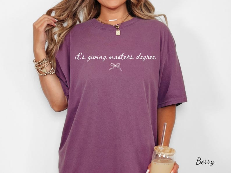 Puede incluir: Camiseta color bayas con la frase "it's giving masters degree" en escritura blanca. Un peque&ntilde;o lazo gr&aacute;fico est&aacute; debajo del texto. La camiseta es de corte holgado y tiene mangas cortas.