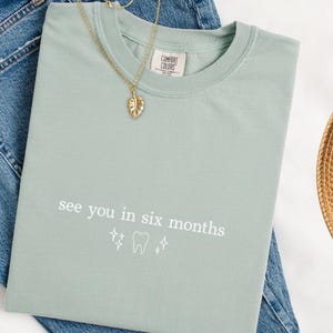 K&ouml;nnte beinhalten: Hellgr&uuml;nes Comfort Colors T-Shirt mit dem Text "see you in six months" und einer Zahn-Grafik. Eine goldene Blatt-Anh&auml;nger-Halskette und blaue Jeans sind ebenfalls im Bild. Ein Strohhut ist sichtbar.