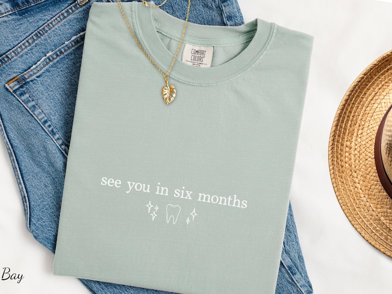 K&ouml;nnte beinhalten: Hellgr&uuml;nes Comfort Colors T-Shirt mit dem Text "see you in six months" und einer Zahn-Grafik. Eine goldene Blatt-Anh&auml;nger-Halskette und blaue Jeans sind ebenfalls im Bild. Ein Strohhut ist sichtbar.