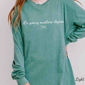 Puede incluir: Camiseta de manga larga verde claro con la frase "it's giving masters degree" en escritura blanca. Un peque&ntilde;o lazo est&aacute; debajo del texto. La camiseta es de color liso y tiene un corte holgado.