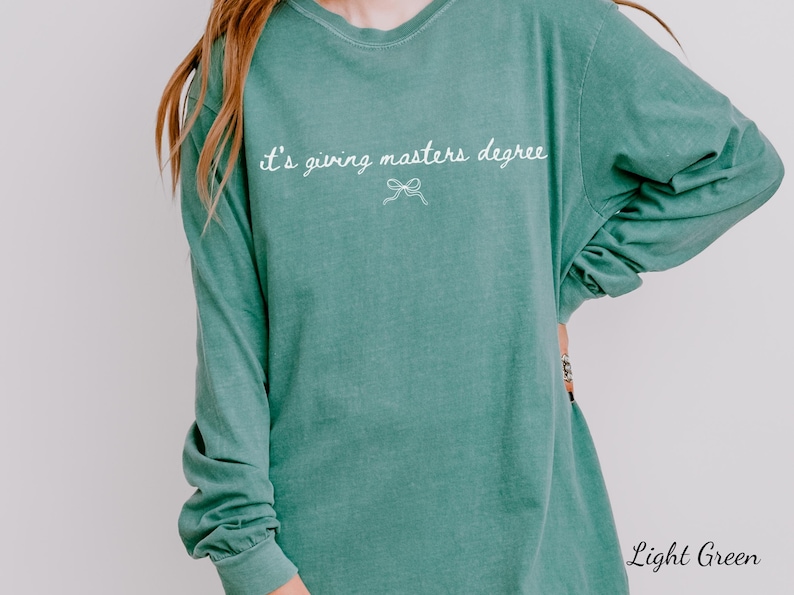 Puede incluir: Camiseta de manga larga verde claro con la frase "it's giving masters degree" en escritura blanca. Un peque&ntilde;o lazo est&aacute; debajo del texto. La camiseta es de color liso y tiene un corte holgado.