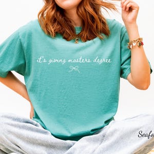 Puede incluir: Camiseta verde mar con la frase "it's giving masters degree" en escritura blanca. Un peque&ntilde;o lazo gr&aacute;fico est&aacute; debajo del texto. La camiseta es de un estilo c&oacute;modo e informal.