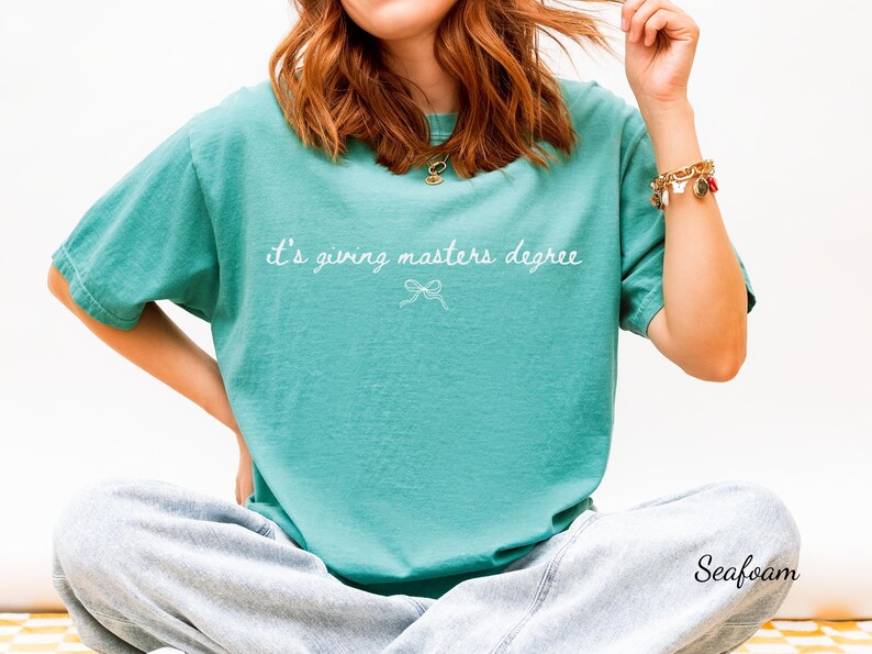 Puede incluir: Camiseta verde mar con la frase "it's giving masters degree" en escritura blanca. Un peque&ntilde;o lazo gr&aacute;fico est&aacute; debajo del texto. La camiseta es de un estilo c&oacute;modo e informal.