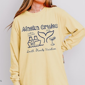 May include: Butter-colored long-sleeve shirt with a navy blue graphic that reads "Alaska Cruise 2026" with a ship, whale tail, and cocktail illustration. The text "making memories" and "Smith Family Vacation" are also included.