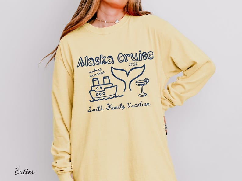 May include: Butter-colored long-sleeve shirt with a navy blue graphic that reads "Alaska Cruise 2026" with a ship, whale tail, and cocktail illustration. The text "making memories" and "Smith Family Vacation" are also included.