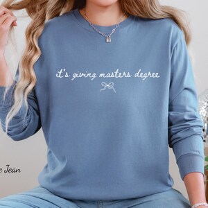 Puede incluir: Sudadera azul de manga larga con el texto blanco "it's giving masters degree" y un peque&ntilde;o lazo. Las palabras "Blue Jean" est&aacute;n impresas en la esquina inferior izquierda.