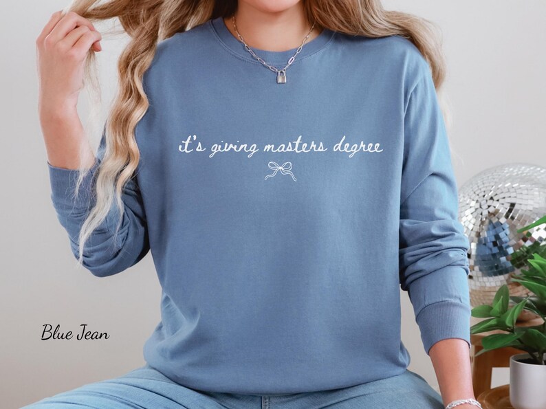 Puede incluir: Sudadera azul de manga larga con el texto blanco "it's giving masters degree" y un peque&ntilde;o lazo. Las palabras "Blue Jean" est&aacute;n impresas en la esquina inferior izquierda.