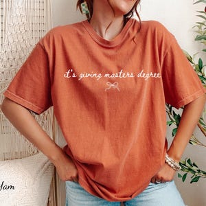 Puede incluir: Camiseta naranja quemado con la frase "it's giving masters degree" en escritura blanca. La camiseta tiene un corte holgado y mangas cortas. El texto est&aacute; centrado sobre un peque&ntilde;o lazo.