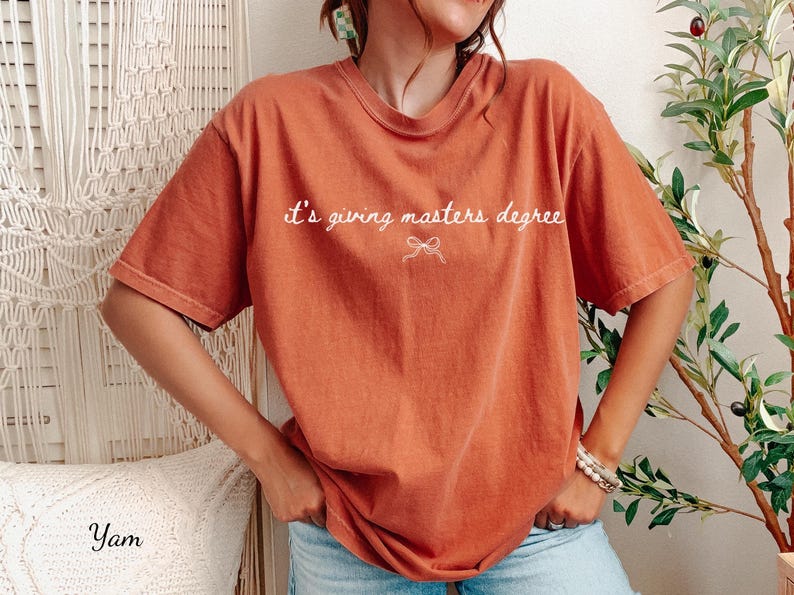 Puede incluir: Camiseta naranja quemado con la frase "it's giving masters degree" en escritura blanca. La camiseta tiene un corte holgado y mangas cortas. El texto est&aacute; centrado sobre un peque&ntilde;o lazo.